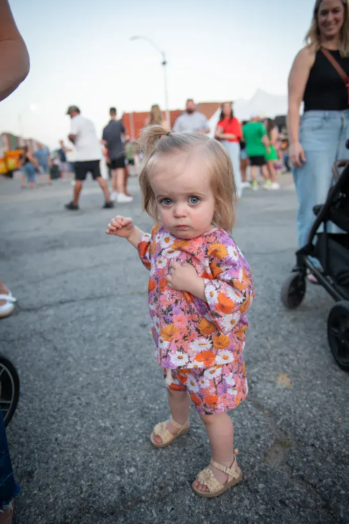 A young toddler in a colorful floral dress stands in the festival grounds with attendees and venue structures visible behind.