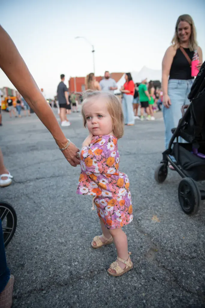 A young child in a colorful floral dress holds an adult's hand while standing in a festival crowd.