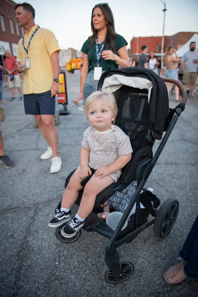 A young child sits in a stroller while festival attendees walk around the outdoor venue in the background.