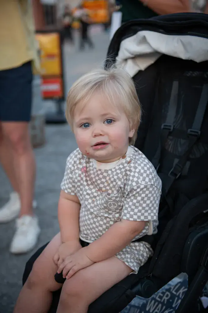A young child in a checkered shirt sits in a stroller at an outdoor festival venue.