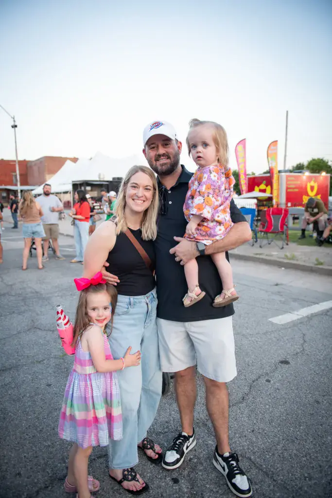 A family of four smiles at the Dancing Rabbit Music Festival with inflatable attractions and crowds visible in the background.