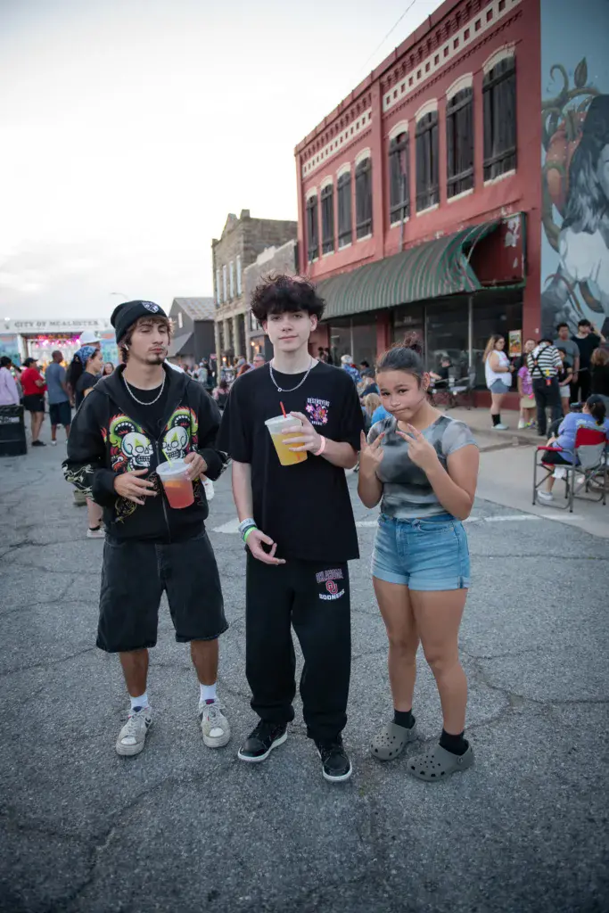 Three attendees holding beverages pose together on a street outside historic red brick buildings at the festival.
