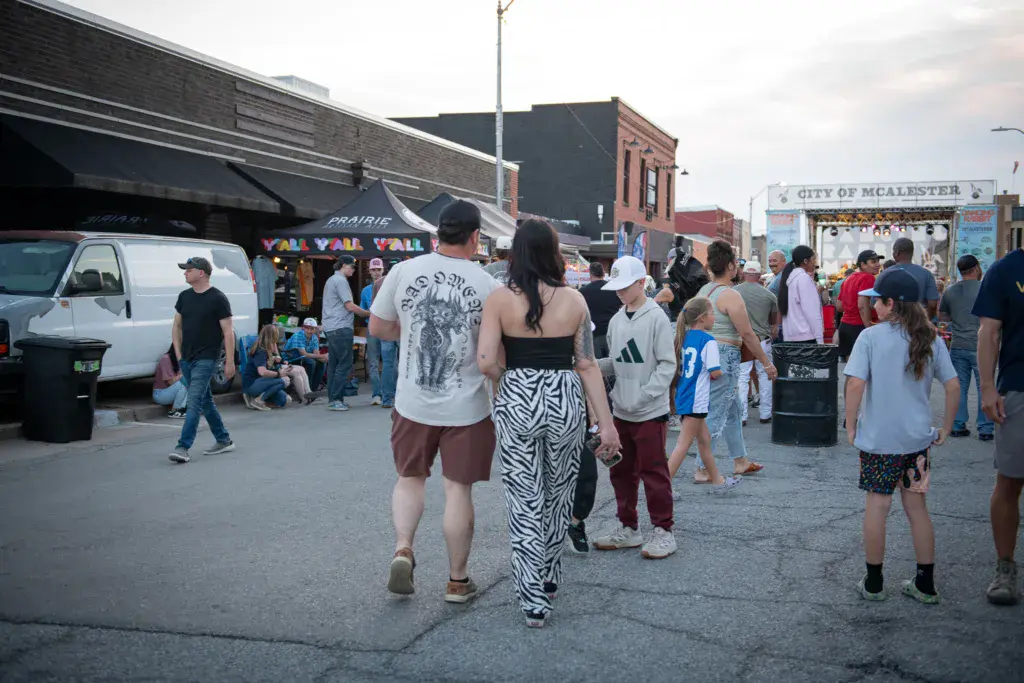 Festival attendees walk through an outdoor venue with urban buildings and a stage structure in the background.