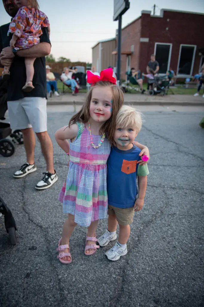 Two young children posing together on a festival street with crowds and brick buildings in the background.