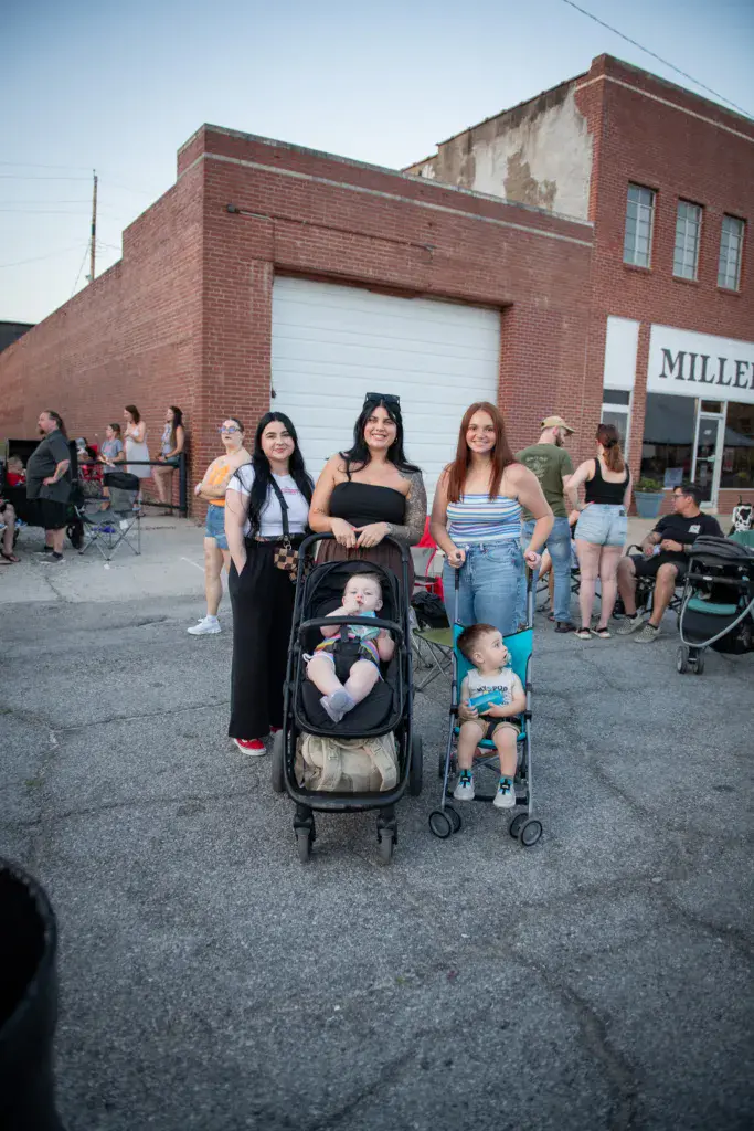 Four women with a baby stroller and toddler stand in a festival crowd outside a brick building.