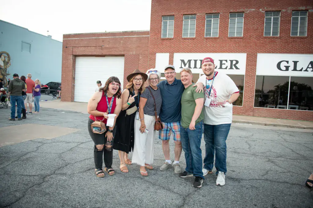 A group of six smiling people pose together for a photo in front of a brick building at an outdoor festival.