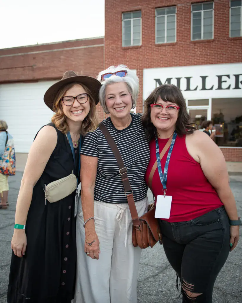 Three women smiling at the camera in front of a brick building at the festival venue.