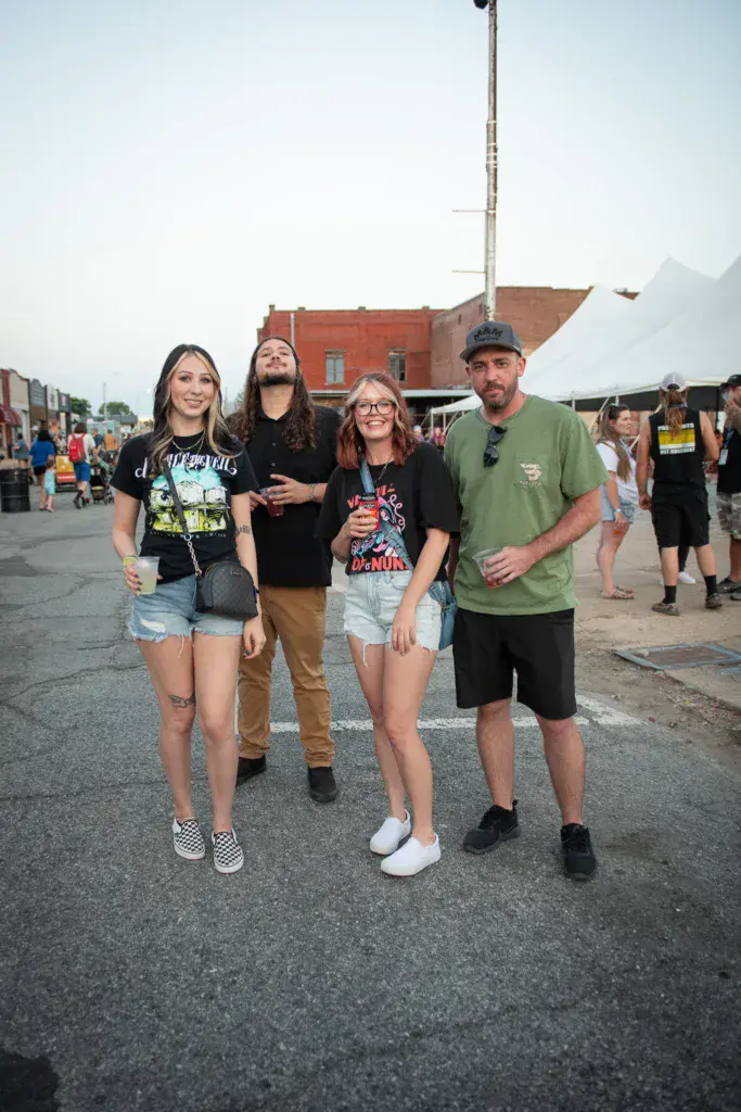 Four festival attendees walking together on a paved area with brick buildings and vendor tents in the background.