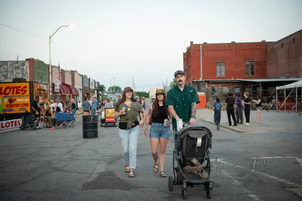 Festival attendees walk through a venue area with food vendors and colorful storefronts visible in the background.