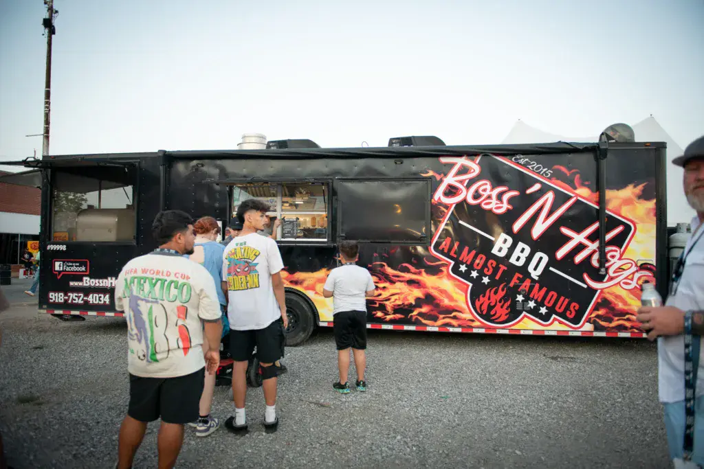 People line up at a food truck with neon signage at an outdoor festival.