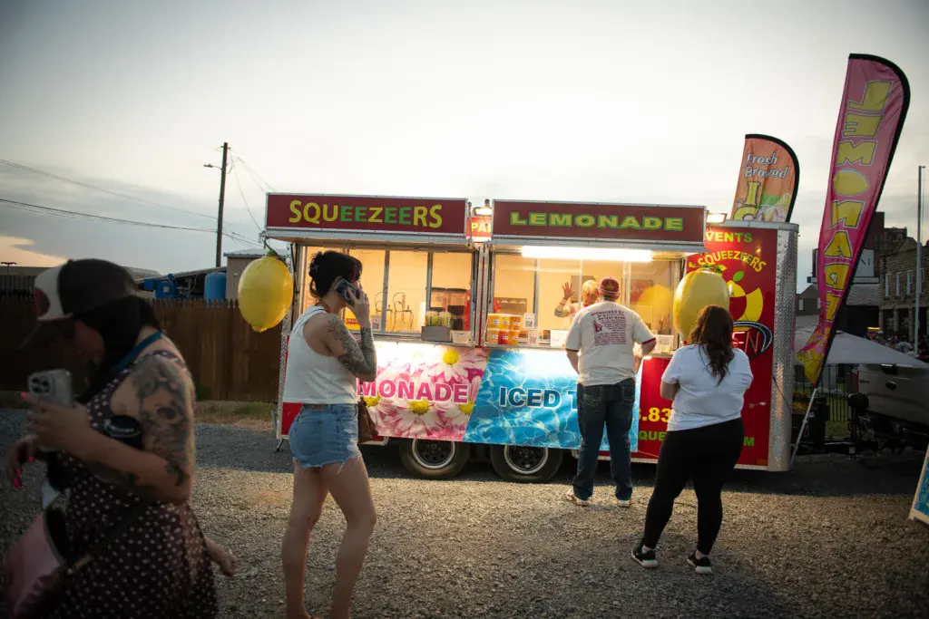 Festival-goers stand near a brightly lit food vendor booth selling lemonade and squeezers at dusk.