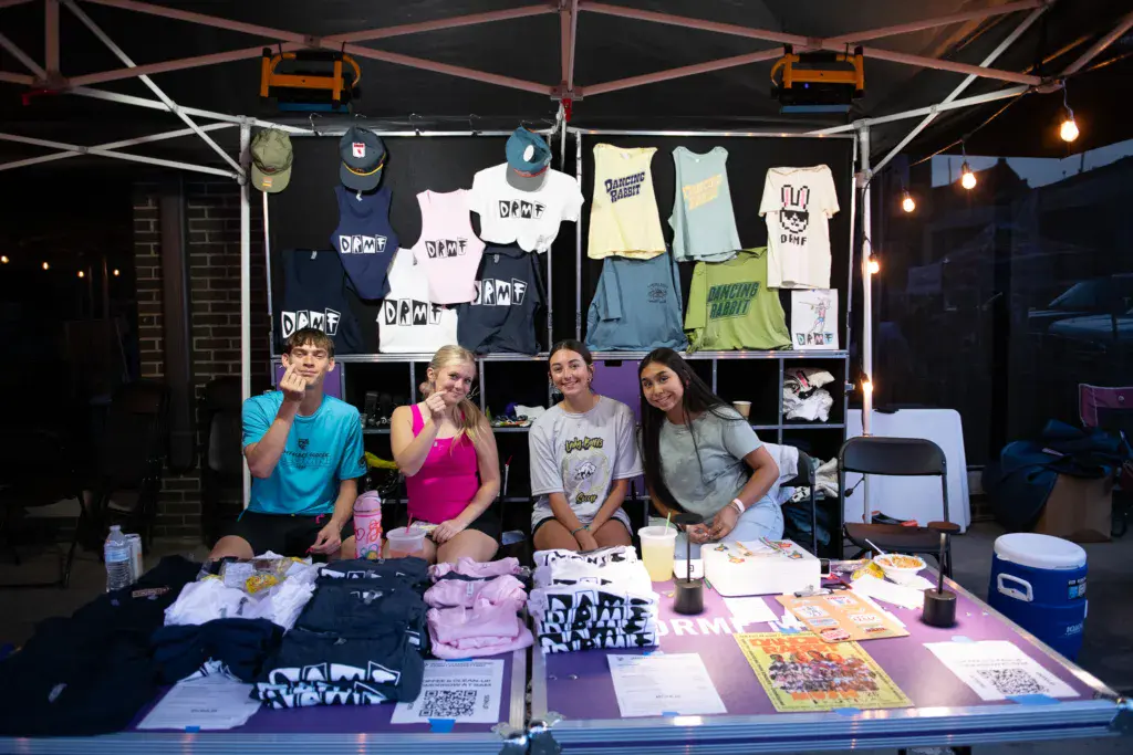 Four people staffing a merchandise tent with branded clothing and promotional materials displayed behind them.