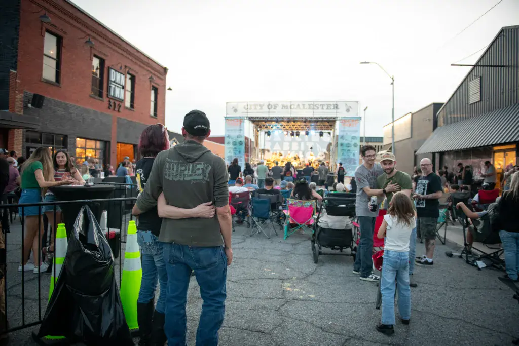 Outdoor festival venue with stage, crowd, and brick buildings in urban setting at dusk.