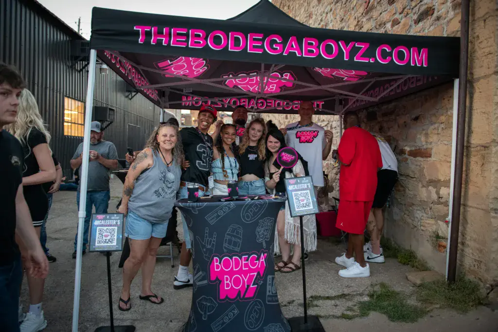 Sponsor booth staff pose together under a black canopy with bright pink neon signage at the festival.