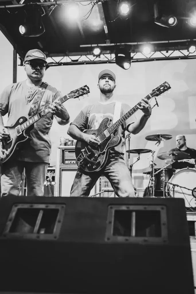 Two guitarists and a drummer perform on an illuminated stage at an outdoor music festival.