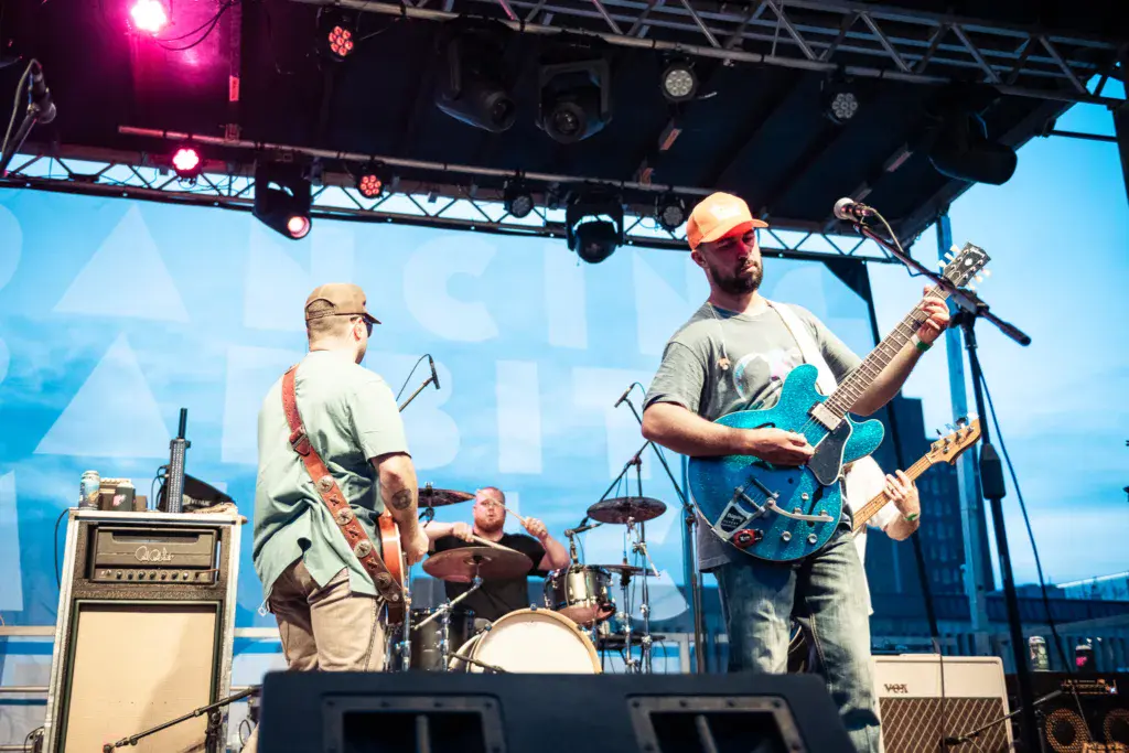 Three musicians perform on stage under bright lighting with blue geometric backdrop and metal truss setup.