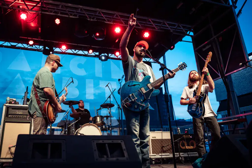 A band performs on stage at sunset with red stage lights, featuring guitarists and a drummer.
