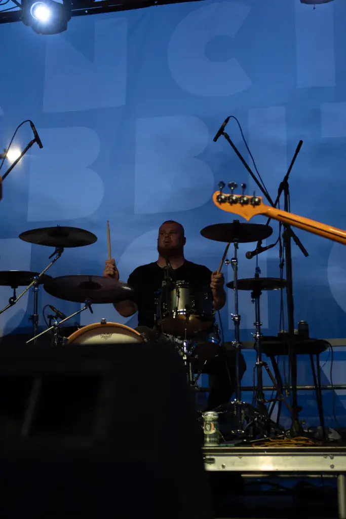 Drummer performing on stage against a blue backdrop with professional stage lighting and equipment
