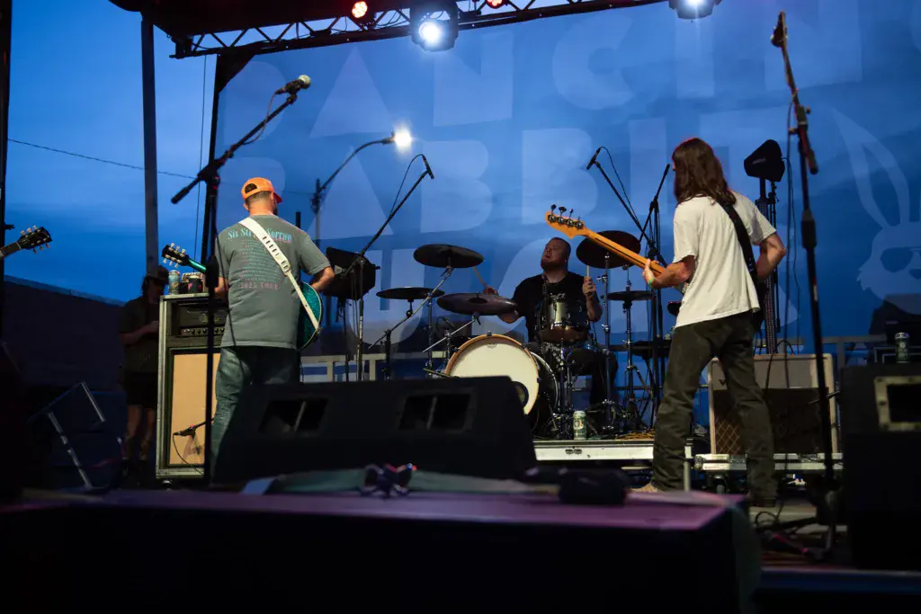A band performs on an illuminated stage at dusk with three musicians visible playing instruments.