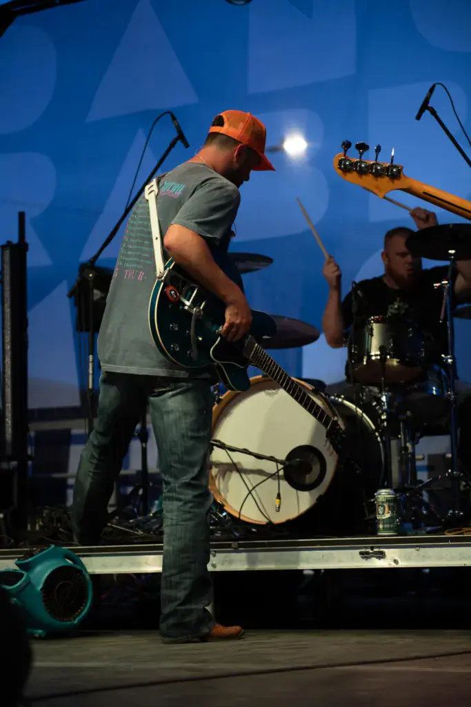 Guitarist wearing an orange cap performs on stage during evening at the festival, with a drummer visible in the background.