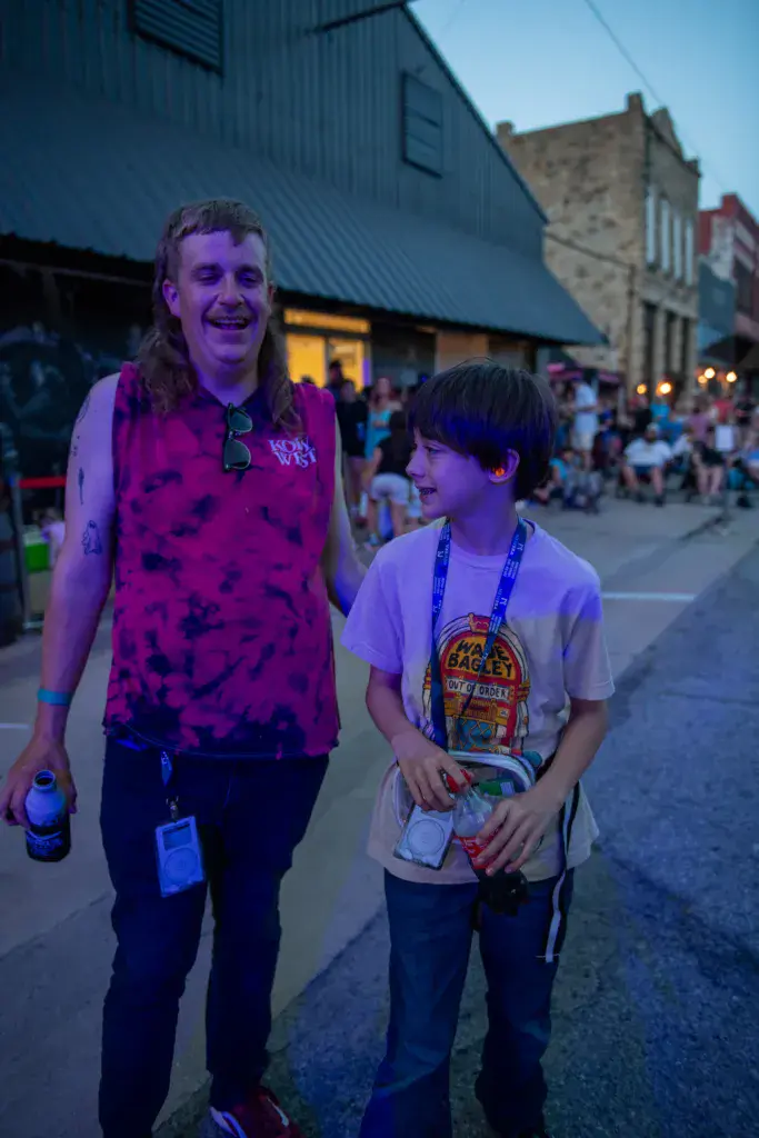 Two festival attendees wearing lanyards walk together through the venue grounds at dusk.
