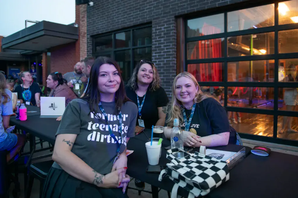 Three people wearing festival lanyards smile at a vendor table outside a brick building at dusk.