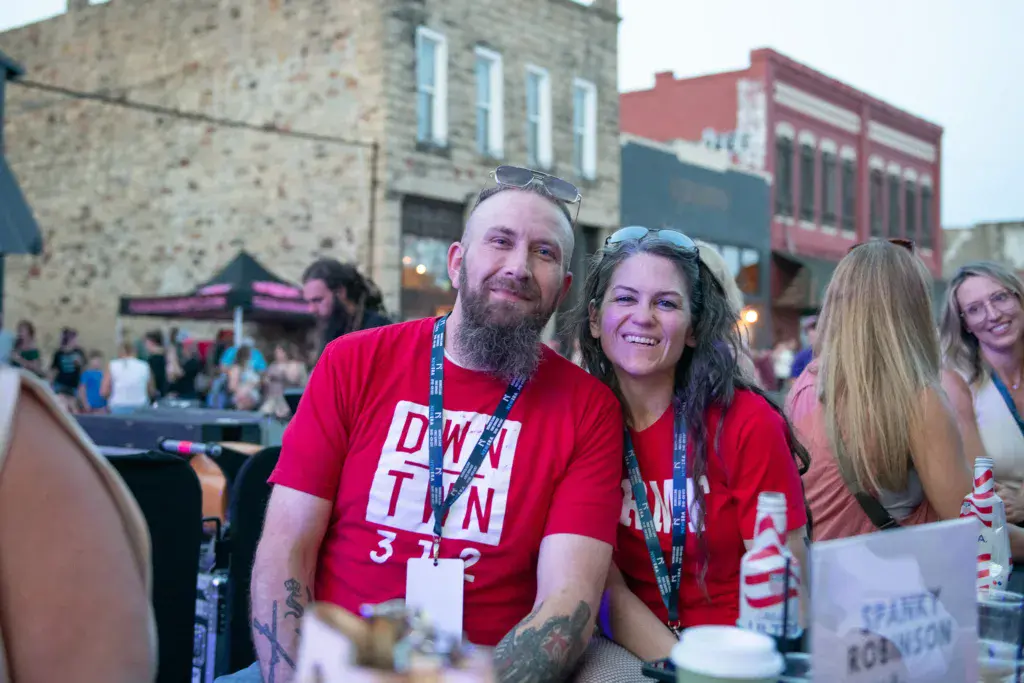 Two people wearing red staff shirts and lanyards smile at an outdoor music festival with historic buildings in the background.