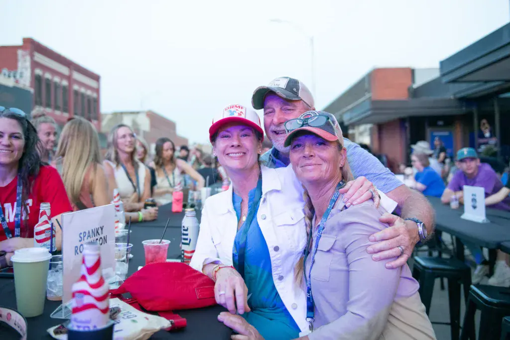 Three people wearing festival apparel smile together at an outdoor table in an urban venue setting.