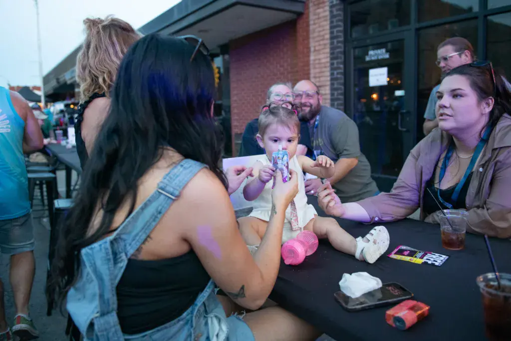 A young child enjoys a treat at a table while surrounded by smiling adults at an outdoor festival venue.
