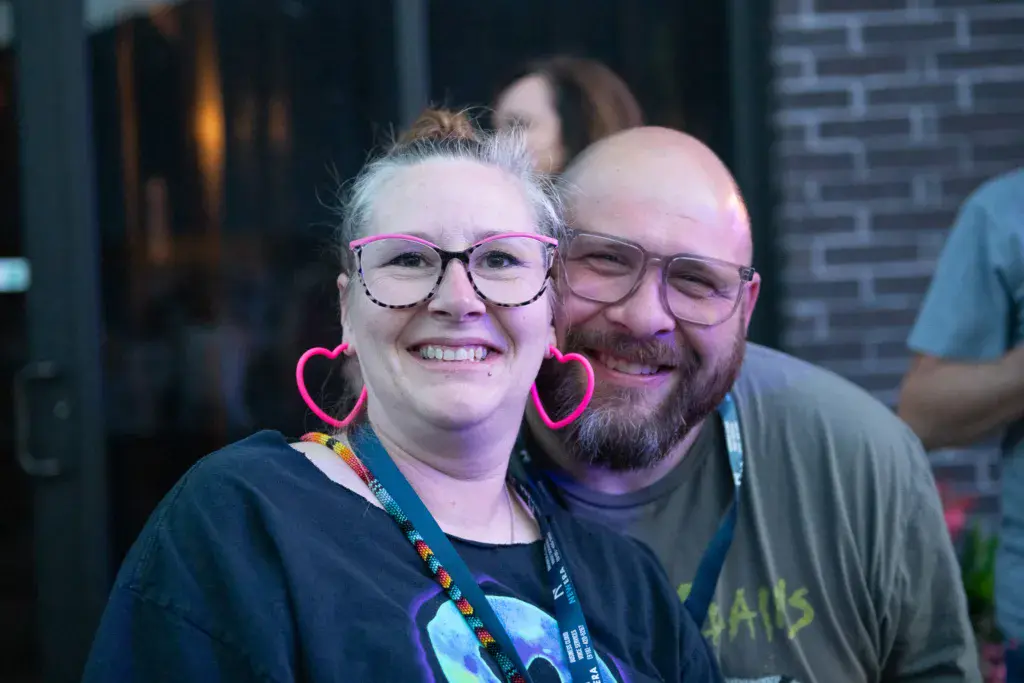Two smiling festival attendees wearing glasses pose together at an indoor venue.