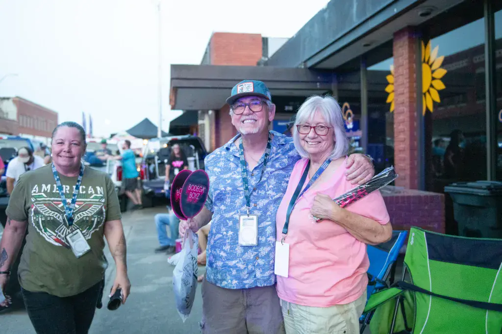 Three smiling festival attendees wearing lanyards stand together on a city street with festival tents and buildings in the background.