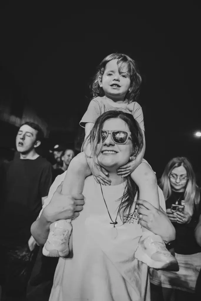 An adult wearing sunglasses holds a young child on their shoulders while standing in a crowd at a nighttime outdoor event.