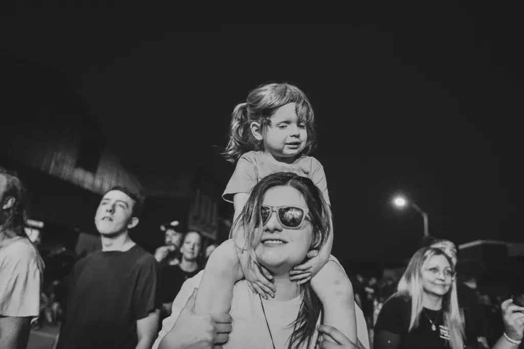 An adult wearing sunglasses holds a young child on their shoulders while standing in a crowd at night.