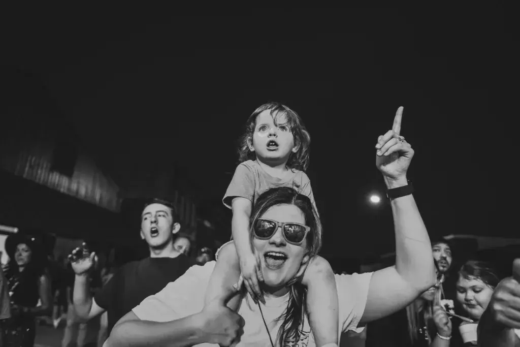 A person wearing sunglasses holds a child on their shoulders while enjoying the music at the festival in black and white.