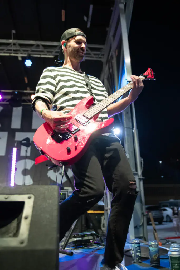 A guitarist wearing a striped shirt performs on stage with a pink electric guitar under stage lighting.