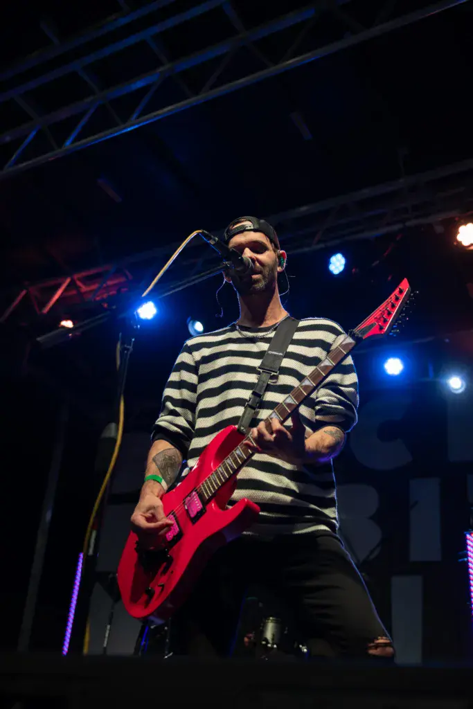 A musician performs on stage with a red electric guitar, wearing a black and white striped shirt under stage lighting.
