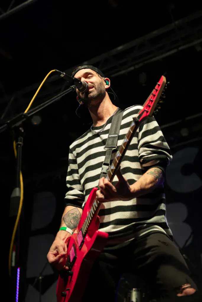 A guitarist and vocalist performs on stage wearing a striped shirt and holding a bright red electric guitar.
