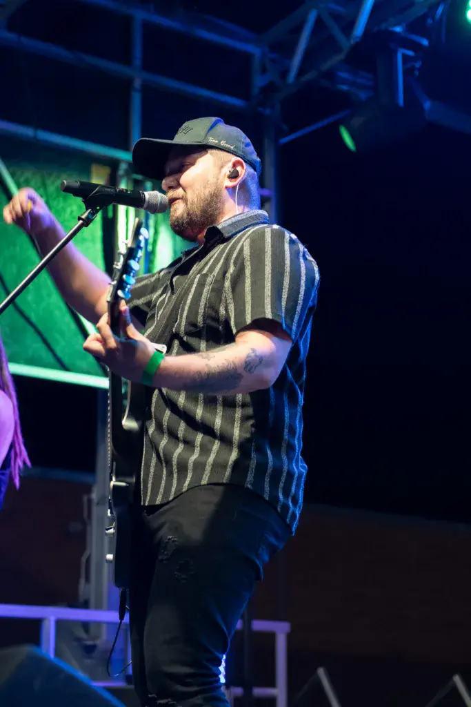 A bearded performer wearing a striped shirt and cap sings into a microphone on an illuminated stage.