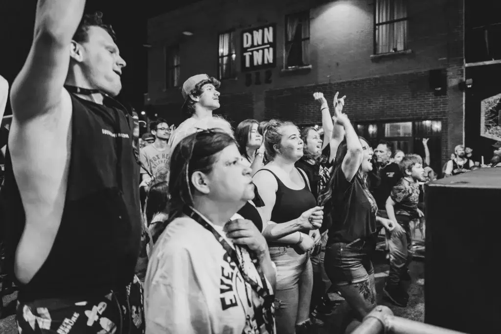 A black and white photograph of festival attendees gathered outdoors, with some raising their arms and engaging with the crowd at night.