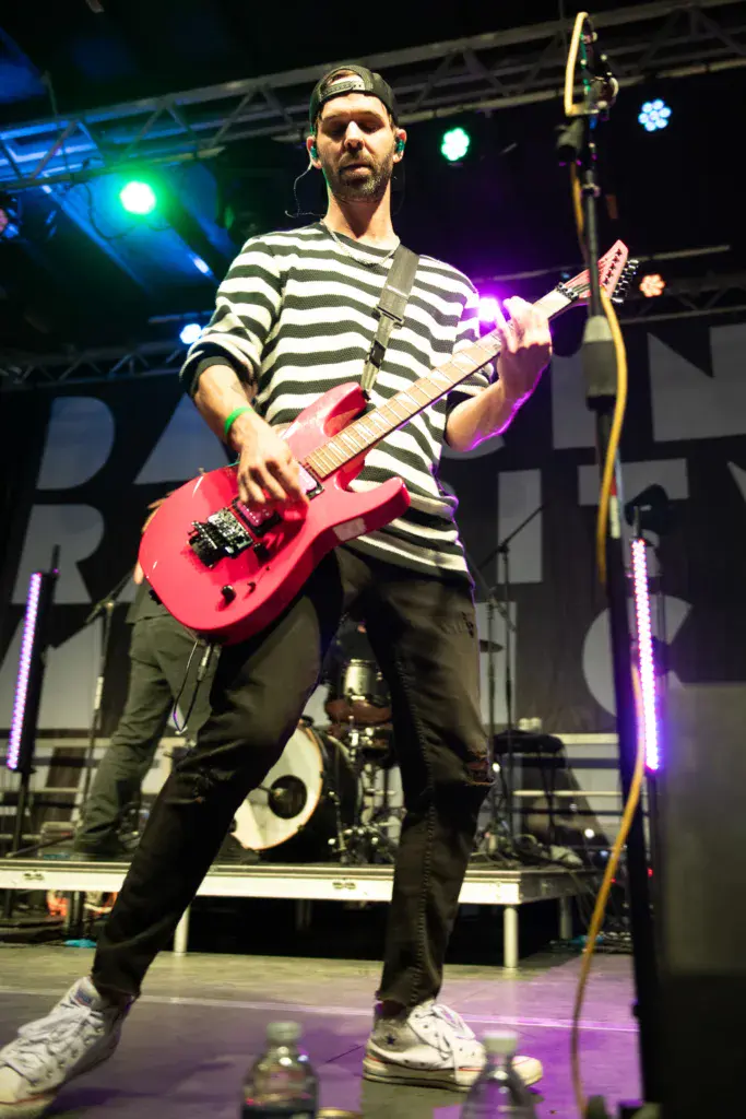 Guitarist in striped shirt performing with pink electric guitar on stage under colorful lights