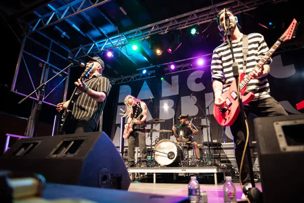 A four-piece band performs on an illuminated stage with colorful lights and a steel truss framework overhead.