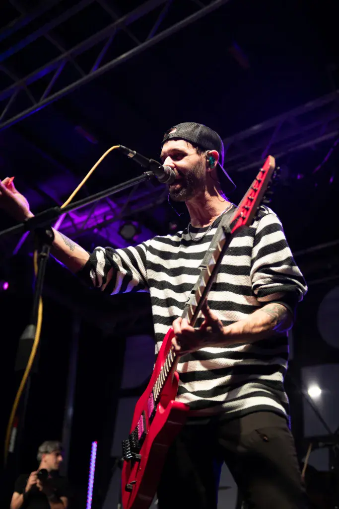 A musician performing on stage with a red electric guitar and microphone under purple stage lighting.