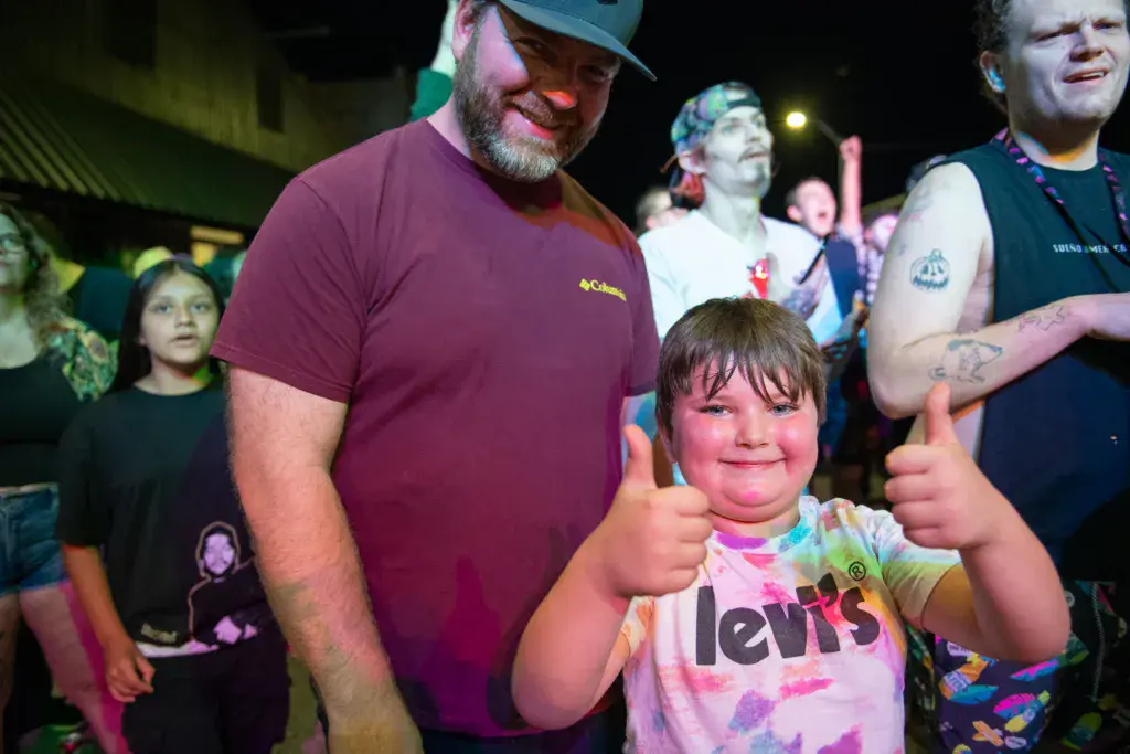 A child gives two thumbs up while celebrating at an outdoor music festival surrounded by festive attendees.