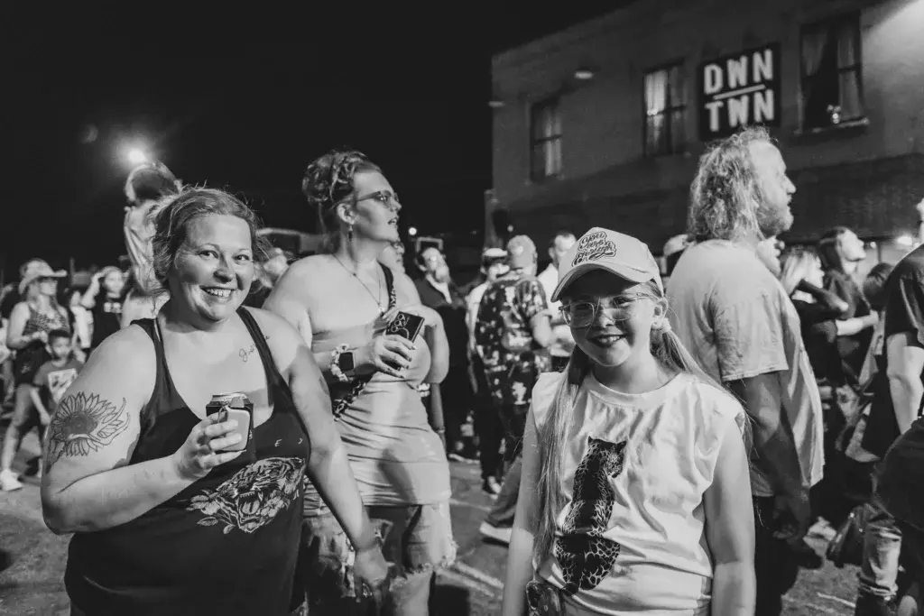 Festival attendees enjoying drinks and smiling at an outdoor nighttime event with venue signage visible in the background.