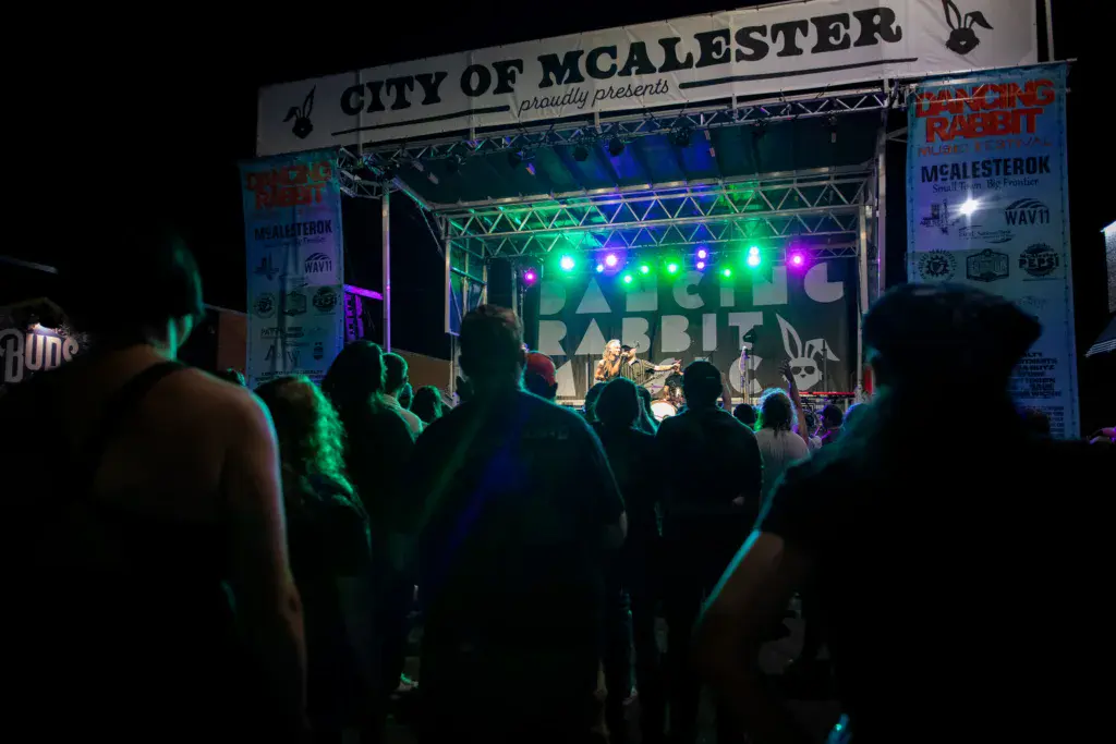 Nighttime festival crowd watches a band perform on an illuminated stage with colorful lights and signage.