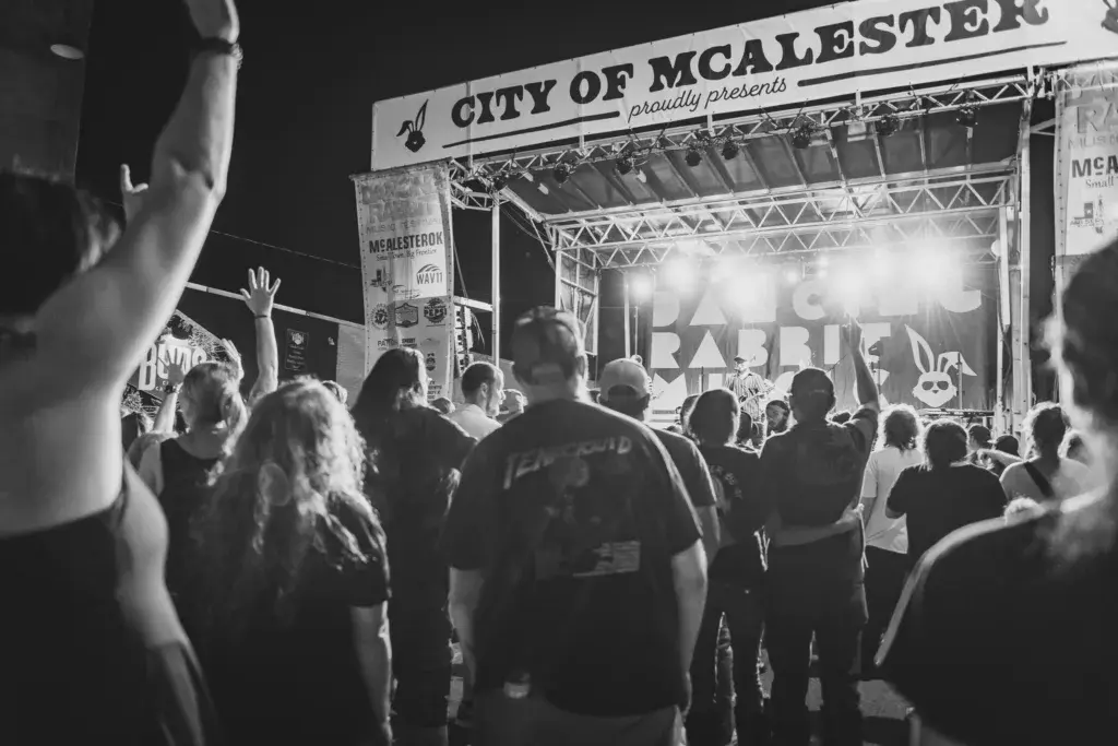 A large crowd of festival-goers faces an illuminated stage with a banner reading 'City of McAlester proudly presents' at a night event.