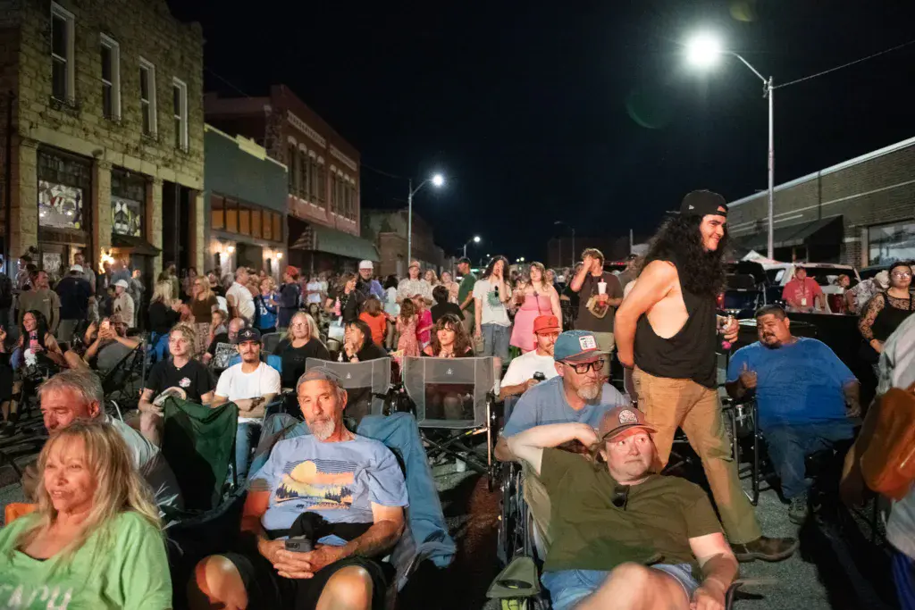 A large nighttime crowd seated in chairs and standing in a town street during an outdoor music festival.