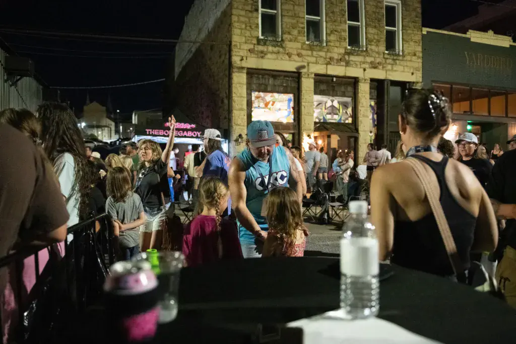 A crowd of people gathering at an outdoor nighttime festival on a city street lined with historic brick buildings.