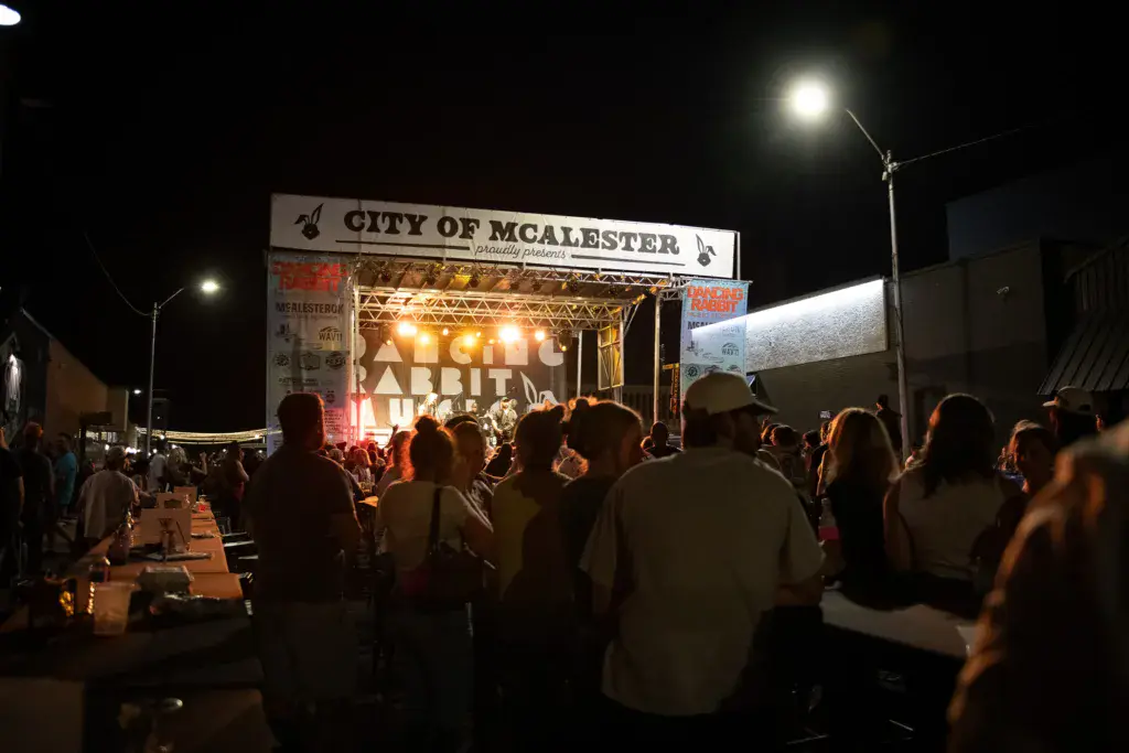 Evening view of a lit stage with crowd gathered in front at an outdoor music festival venue.