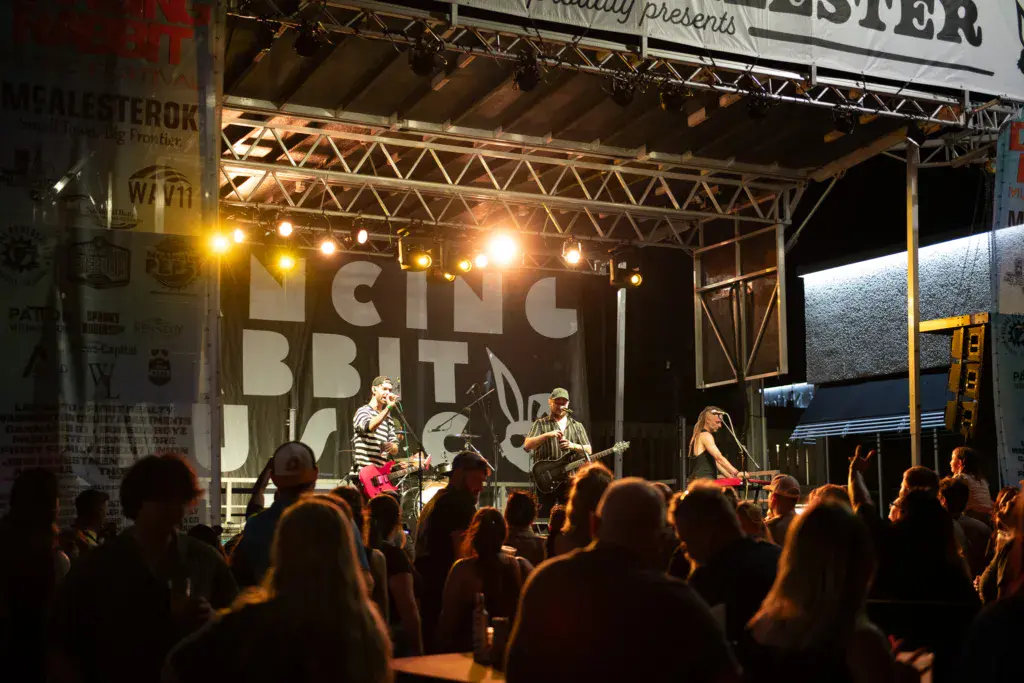 A band performs on an illuminated stage before a crowd of festival attendees at dusk.